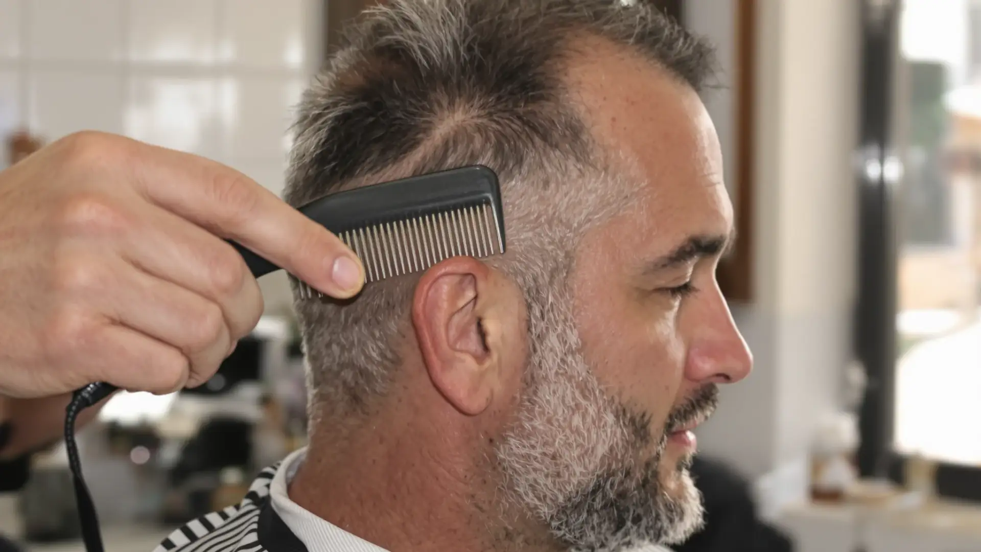 Barbiere che pettina i capelli di un uomo di profilo, con l'attaccatura frontale ben visibile.