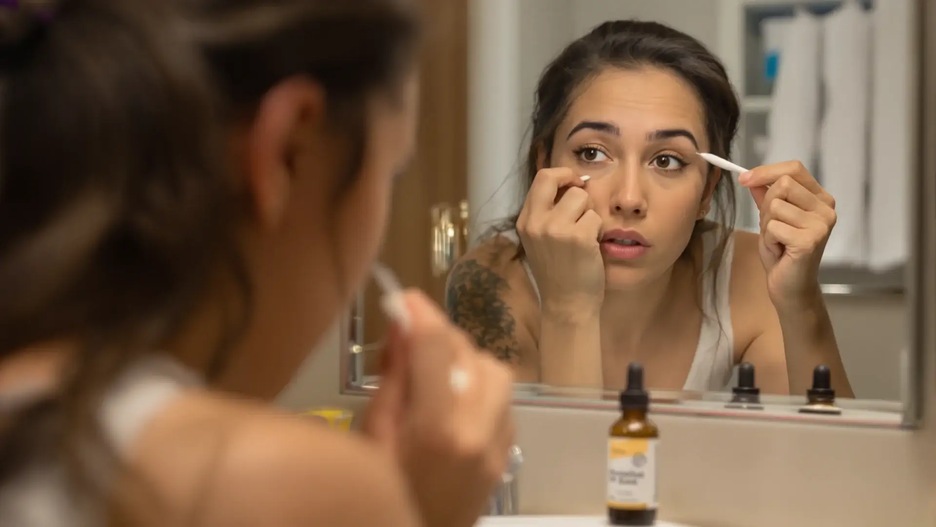 Woman applying minoxidil to her eyebrow with a cotton swab in a bathroom mirror