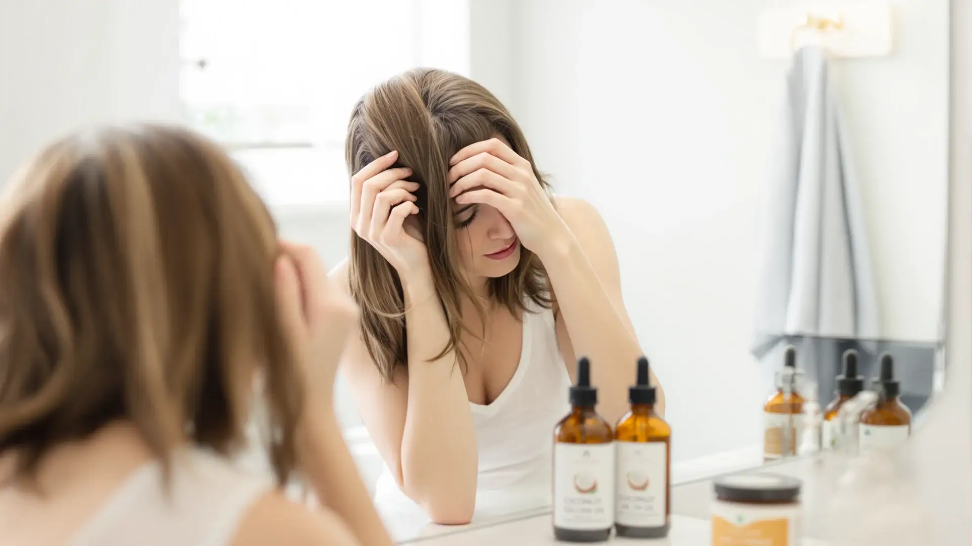 Woman checking her scalp in a bathroom mirror, oregano oil bottle nearby