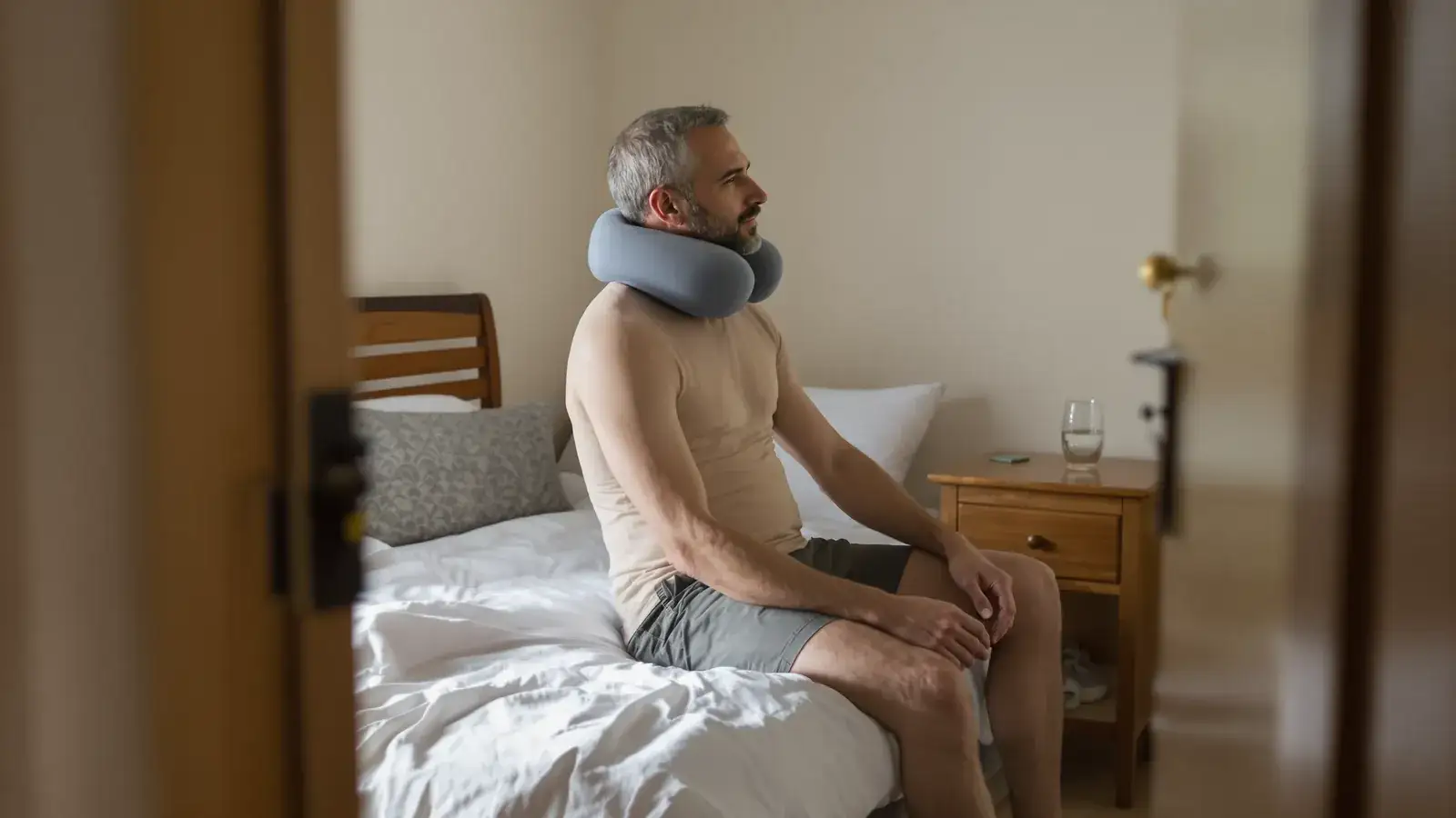 Uomo seduto sul bordo del letto con un cuscino cervicale, in fase di riposo post-operatorio.