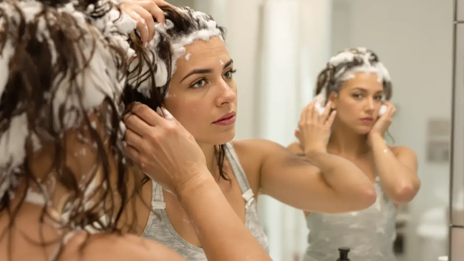 Woman applying scalp treatment to thinning hair in a bathroom mirror.